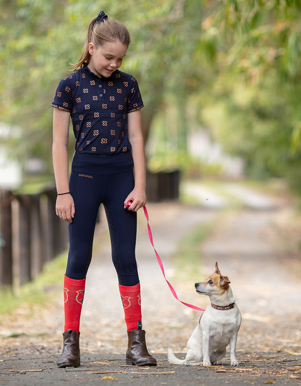 This image shows a young girl wearing a navy and gold riding top and horse riding tights paired with red riding socks. She has a dog sitting beside her feet.