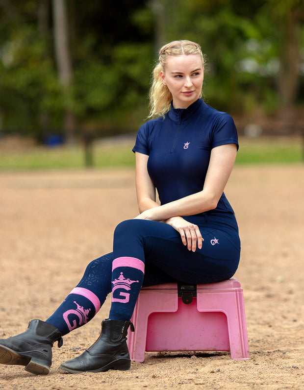 A young, blonde woman in a navy horse riding top and riding tights sits on a pink mounting block.